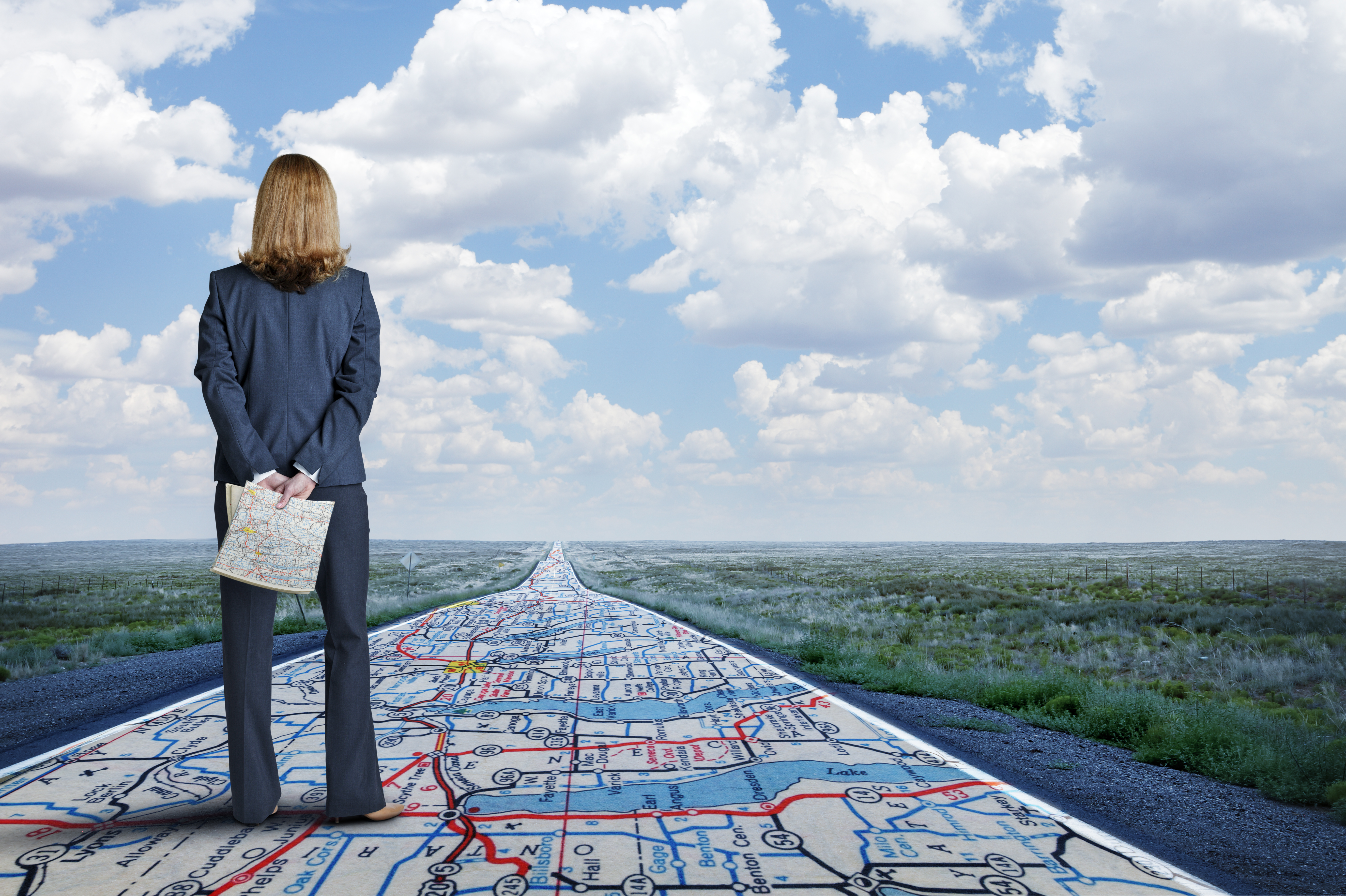 A businesswoman stands while holding a road map in her hands as she looks down a long, straight, rural road that has the same road map painted on it.